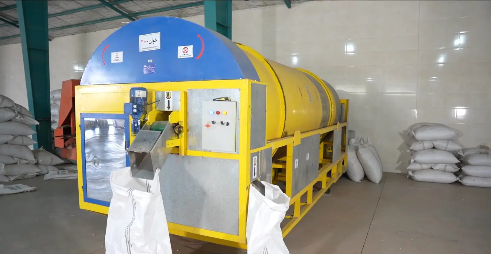 Yellow rotary drum on the processing line, with white bags beside it, in the Aria Saramad factory